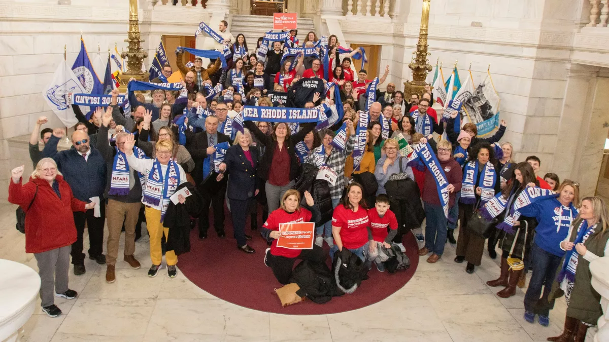 NEARI members pose for a group photo on the interior State House steps on Lobby Day 2024. They are holding up scarves that have the NEARI logo and smiling.