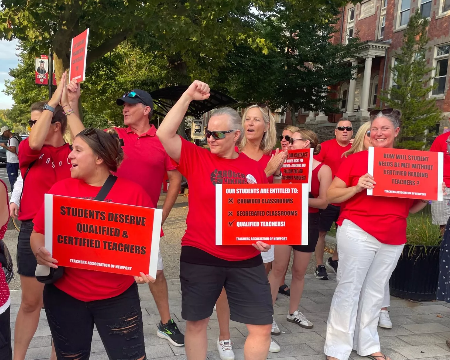 Educators dressed in red on a picket line look to the road with fists up and arms flexed.